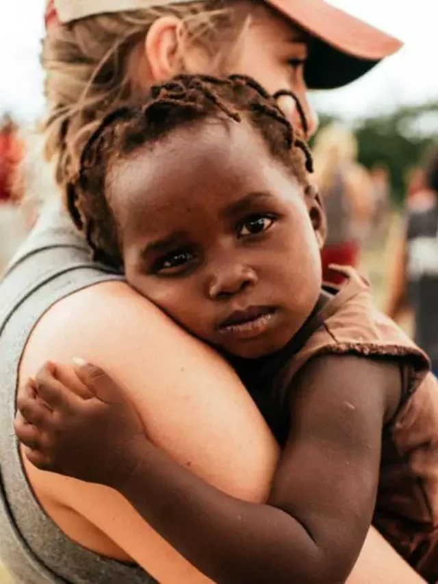 Uma mulher branca abrançando uma criança negra de cerca de quatro anos, aparentemente de uma família com renda muito baixa.